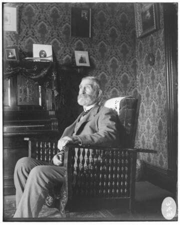 An elderly bearded man sitting in an arm chair.  He is in a residence. surrounded by a piano. a trumpet. portrait photographs. and wall-