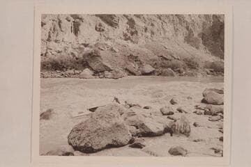 Lining of the Nevills skiffs at Lava Falls. Nevills is standing in the skiff. Joe Desloge is helping in shoving the skiff over the rocks. Kent Frost is snubbing among the rocks at right center