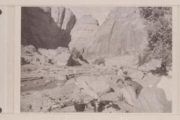 Camp in Navajo Canyon at Pothole Bridge. Ballard Atherton helps Tom Daly at the kitchen. Bahe sits in the background. The creek cuts through left center