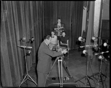 Cinematographer filming two ladies seated on stage.  Man holding slate in back of them says:  "Its So Easy"