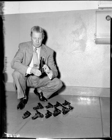 Man kneeling holding automatic pistol with a variety of pistols (revolvers, automatics and a derringer) spread out on the floor beside him