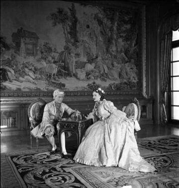Couple in period costume seated at a small table in the library of the Huntington residence with a Beauvais tapestry in the background