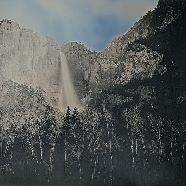 A waterfall cuts through a crevice in a tall mountain range, disappearing behind a forest of trees.