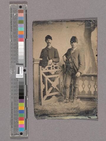 Portrait of two unidentified men posed at a gate with a studio backdrop