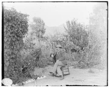 A man sitting in a rocking chair in the garden of a residence. same shot as 01-